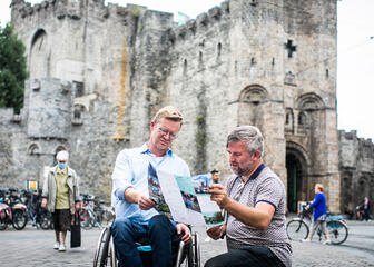 een man die een kaart toont aan een andere man in een rolstoel, voor het gravensteen