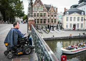 een man in een rolstoel op de st-michielsbrug die uitkijkt over het water, een boot met mensen op het water
